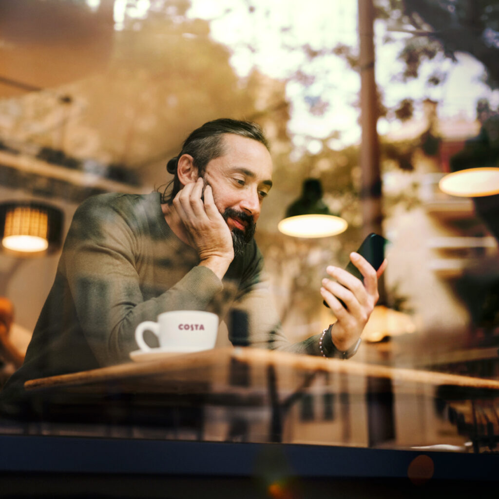 Man sitting at a café table holding a smartphone, with a Costa Coffee cup in the foreground